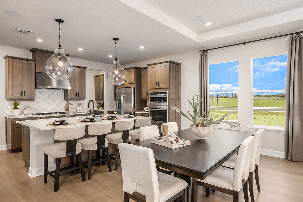 Modern kitchen and dining area with beige chairs, dark wood table, island with bar stools, pendant lights, stainless steel appliances, and large windows showing a scenic outdoor view.