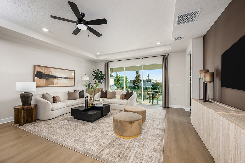 Modern living room with a large beige sectional sofa, brown and gold ottomans, a black coffee table, a neutral area rug, wall art, a ceiling fan, TV, and sliding glass doors opening to a patio with outdoor seating.