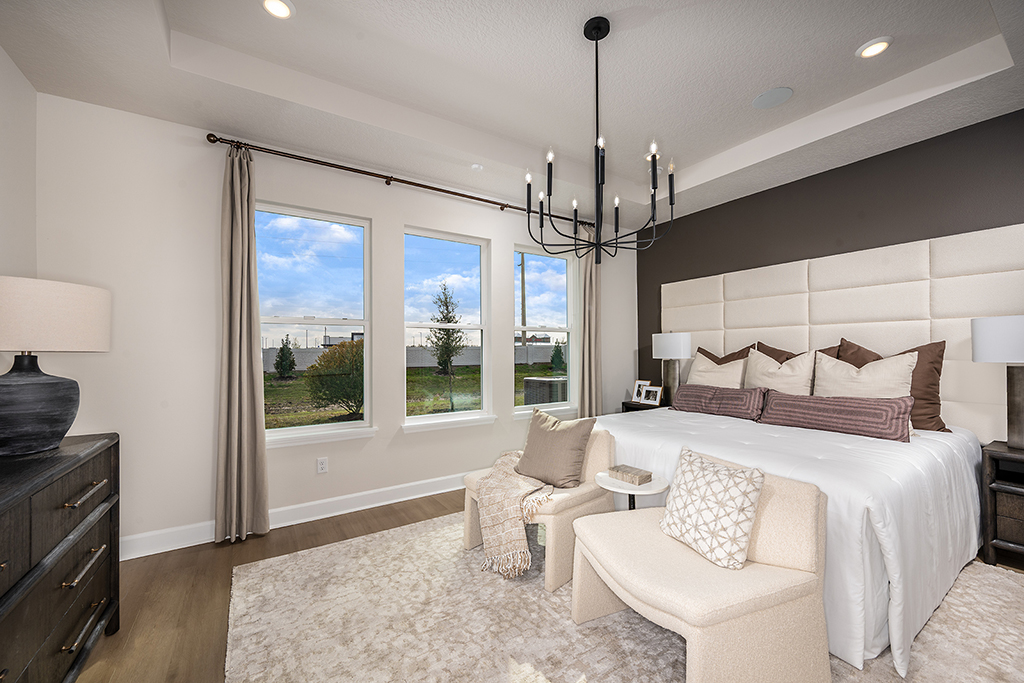 A modern bedroom with a large bed, beige and brown decor, two chairs with a small table at the foot of the bed, a dark dresser, a chandelier, and three large windows letting in natural light.