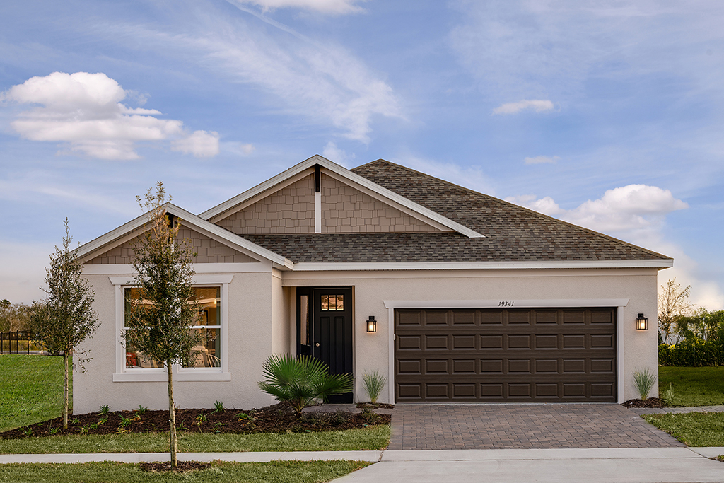 Single-story suburban house with beige siding, brown shingle roof, dark brown garage door, central entrance, two small trees, and minimal landscaping, set against a blue sky with scattered clouds.