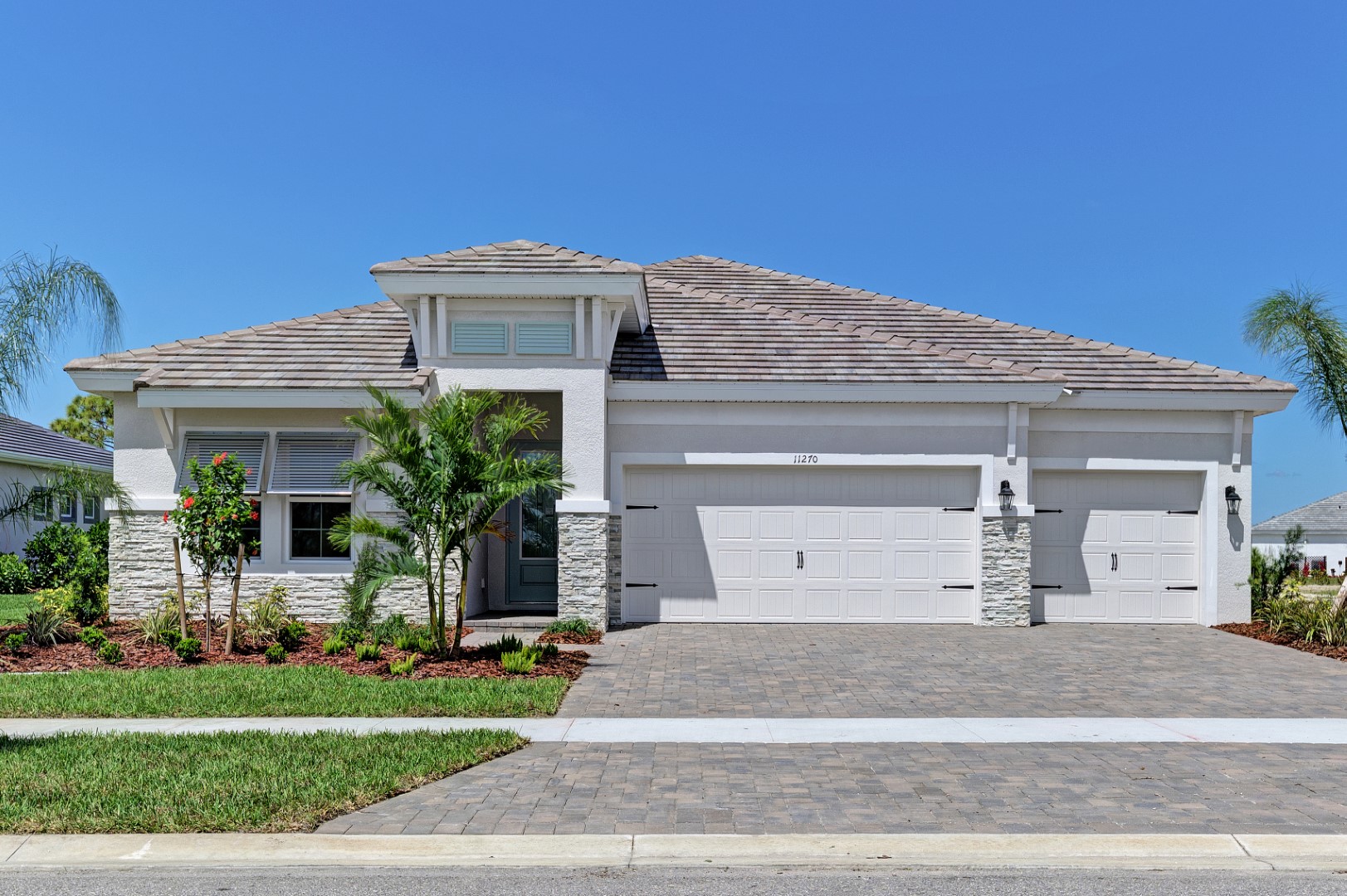 Single-story modern house with a light-colored exterior, three-car garage, tiled roof, manicured lawn, landscaped garden, and palm trees under a clear blue sky.