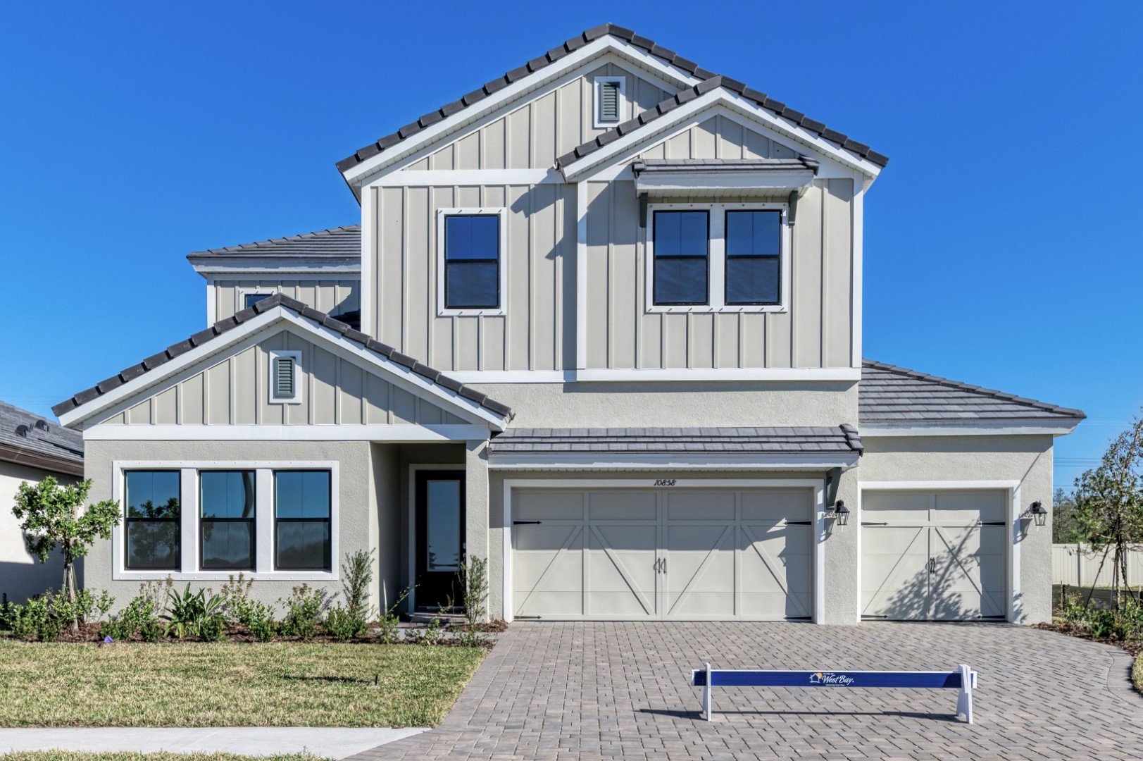 A modern two-story house with light-colored siding, multiple gables, a three-car garage, and a brick driveway, set against a clear blue sky. A blue real estate sign stands in front of the house.