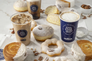 Various pastries, including donuts, croissants, and a pecan-topped pastry, are displayed with iced and hot coffee drinks in Paris Baguette cups. Some pastries are dusted with powdered sugar and nuts, all set on a white surface.