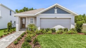 Single-story suburban house with light gray siding, white trim, a two-car garage, and a paved walkway bordered by shrubs and green grass, set on a sunny day with a clear blue sky.