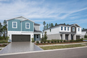 Two modern two-story suburban houses with gray driveways, manicured lawns, and small shrubs in front. The sky is clear with a few clouds, and tall trees are visible in the background.