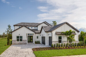 Modern, two-story house with light-colored siding, stone accents, and a dark tiled roof. The front features large windows, a small porch, and a two-car garage. Well-manicured lawn and palm trees surround the paved driveway.