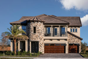 Two-story modern house with a stone facade, arched entryway, round turret, balcony, wooden double garage doors, palm tree, and clear blue sky in the background.