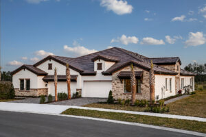 Single-story modern suburban house with a brown tile roof, stone and stucco exterior, three garages, landscaped front yard, palm trees, and a blue sky with clouds in the background.